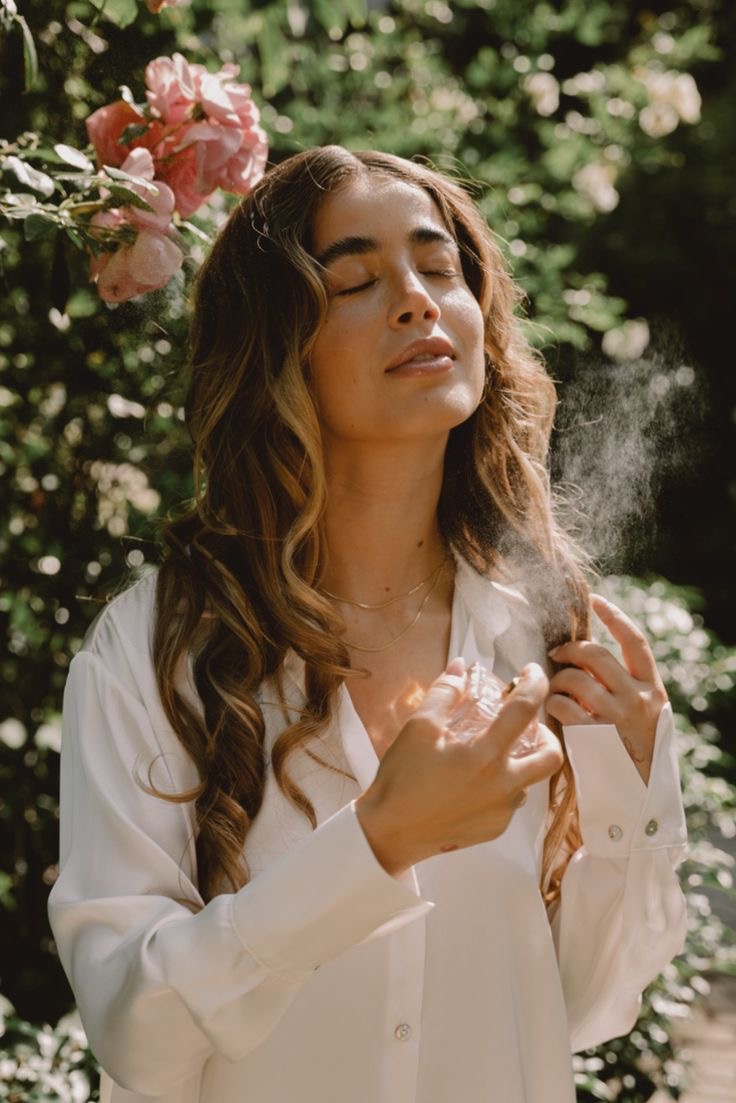 An image of a woman in a white button-up spritzing perfume on her neck and strands of hair.